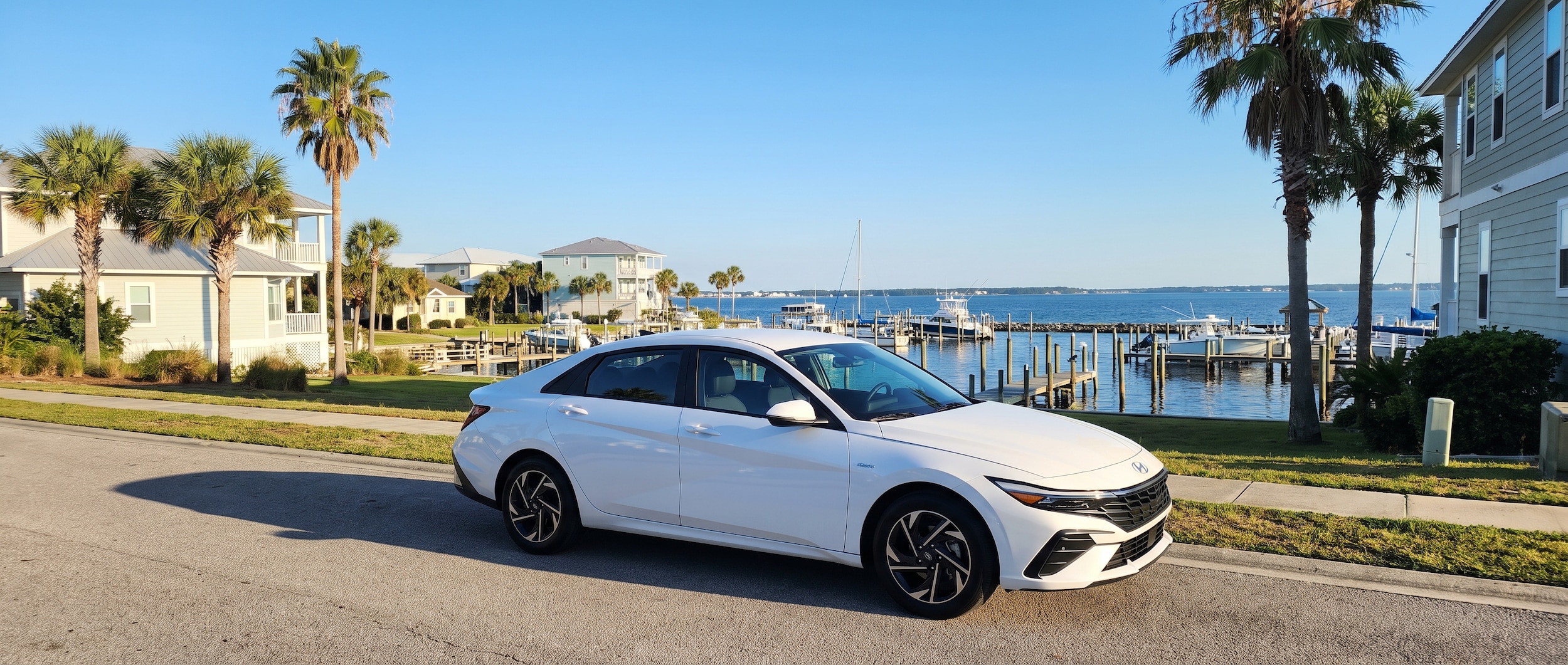 A 2026 Hyundai Elantra Hybrid in a coastal Gulf Coast setting, parked on a sun-drenched street near a waterfront neighborhood, blue summer sky overhead and a bayfront marina visible in the background, Panama City FL