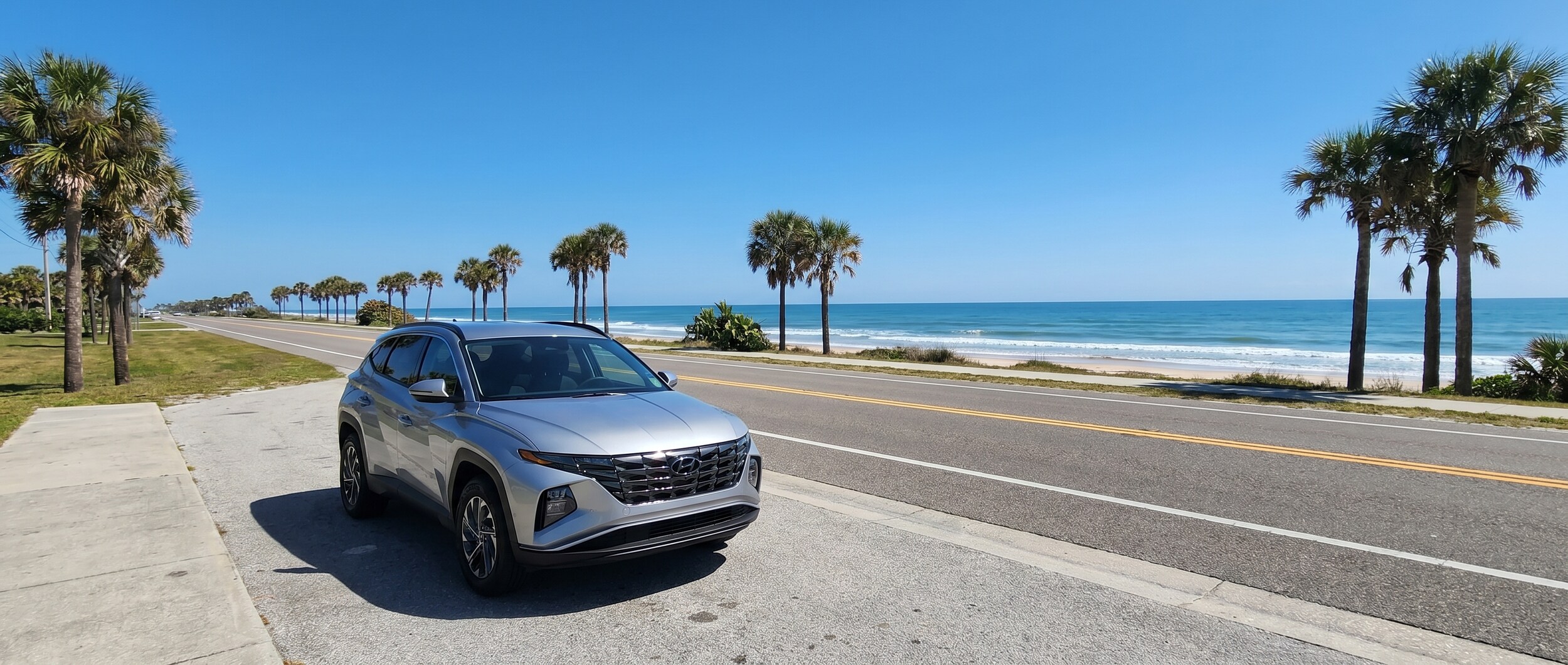 A silver Hyundai Tucson parked along a scenic Gulf Coast highway with clear blue skies, palm trees, and the Florida coastline visible in the background, suggesting a relaxed coastal road trip