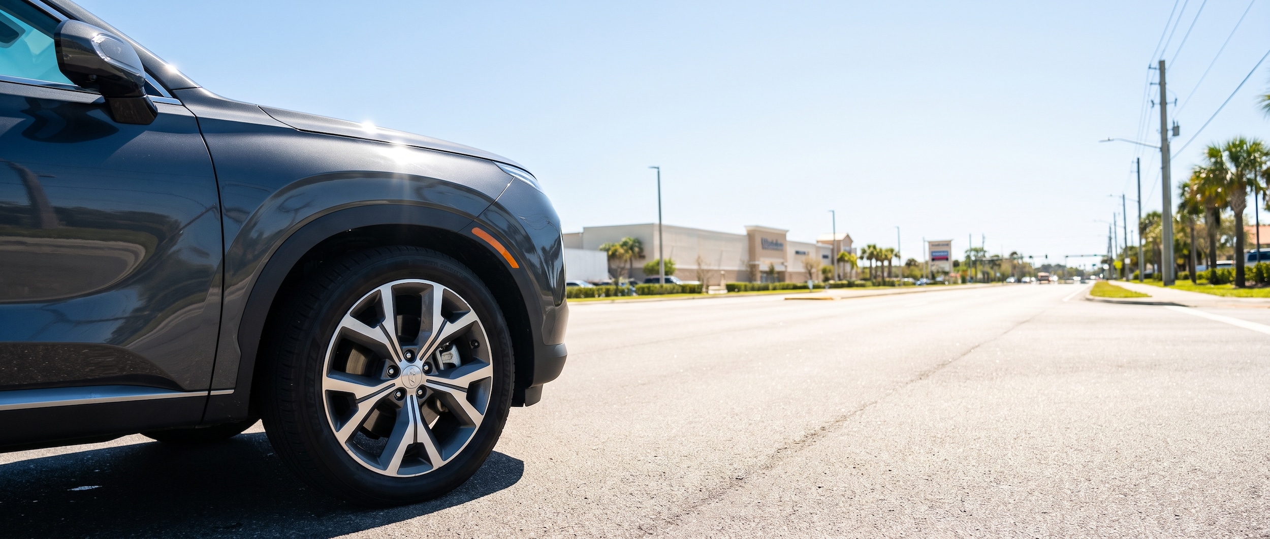 A Hyundai SUV stopped at a traffic light on a sunny, heat-shimmering commercial corridor lined with shopping centers -- wide four-lane road, blue sky, Florida summer light, tire and brake focus implied by a low-angle shot near the wheel well.