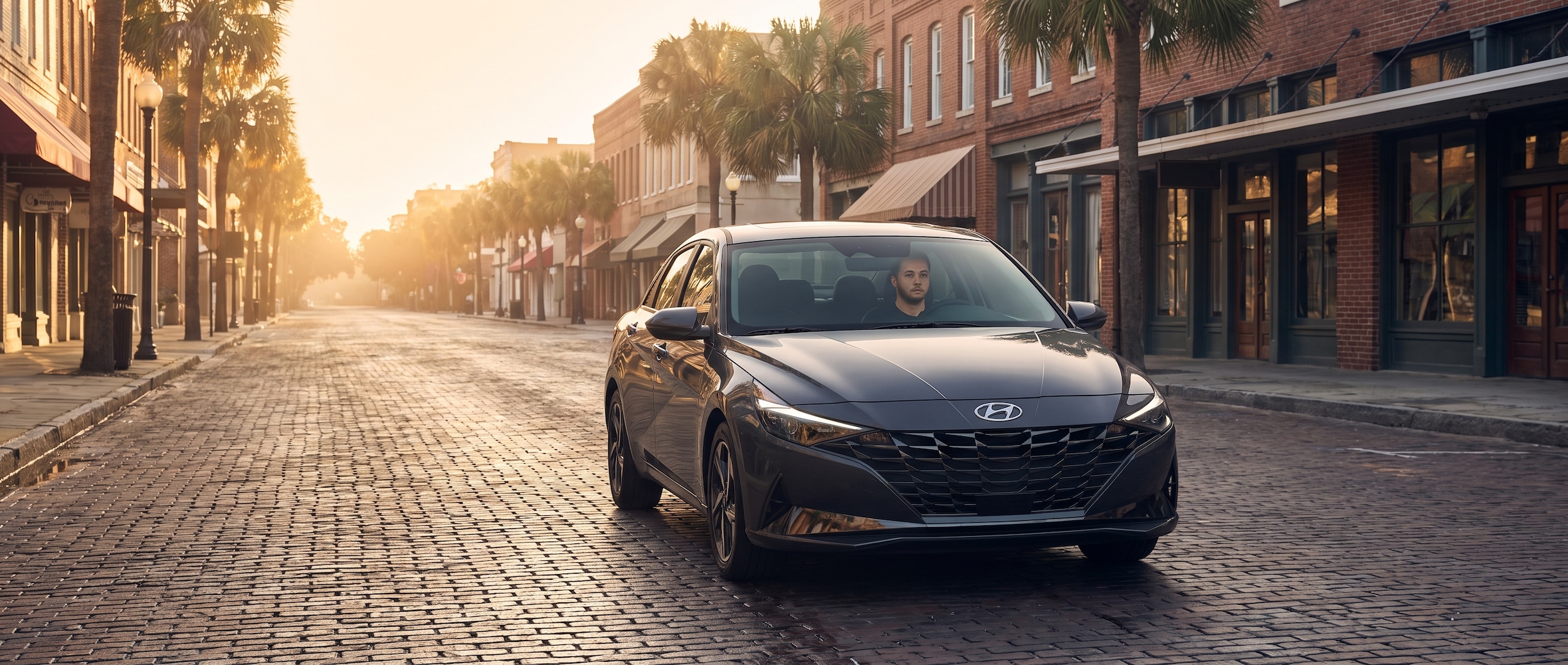 A 2026 Hyundai Elantra Hybrid in a sleek dark color moving through a wide, brick-lined downtown street bathed in early morning Gulf Coast light, palm trees and low-rise storefronts visible, warm Florida sunrise tone