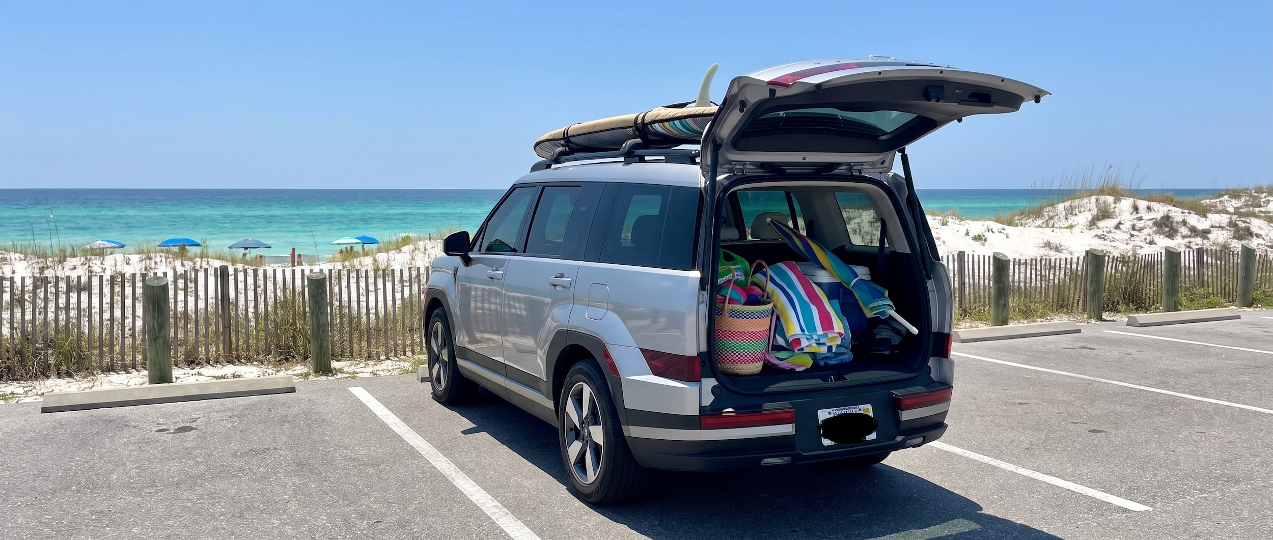 A silver or blue 2025 Hyundai Santa Fe Hybrid SUV loaded with beach gear -- surfboard visible in roof rails, bright Florida summer light, parking lot near a Gulf Coast state park with white sand dunes and emerald water visible in the background. Rear hatch open with colorful beach bags and towels inside.