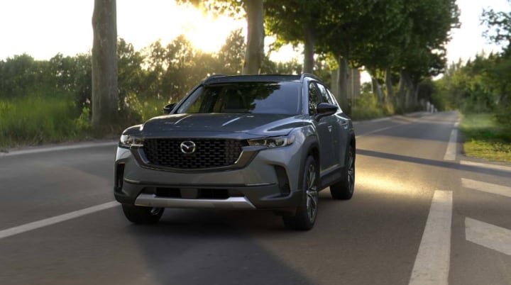 A 2026 Mazda CX-50 driving on a tree-lined road at sunset.