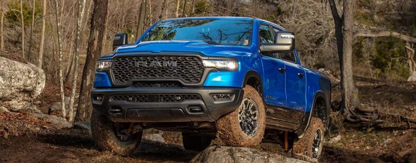A blue 2025 Ram 1500 Rebel off-roading over muddy rocks.