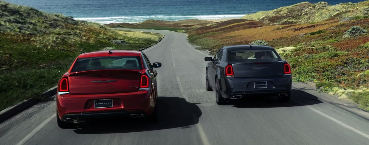 A red and a grey Chrysler 300 are shown from the rear on a coastal road after leaving a Chrysler dealership.
