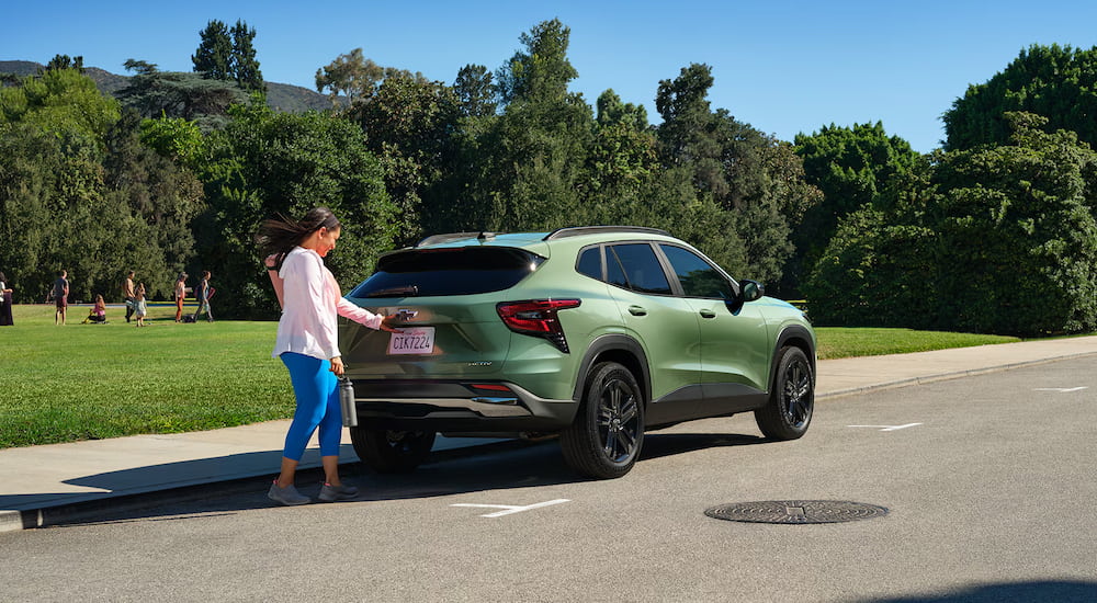 A woman accessing the trunk of a green 2025 Chevy Trax ACTIV parked near a field.