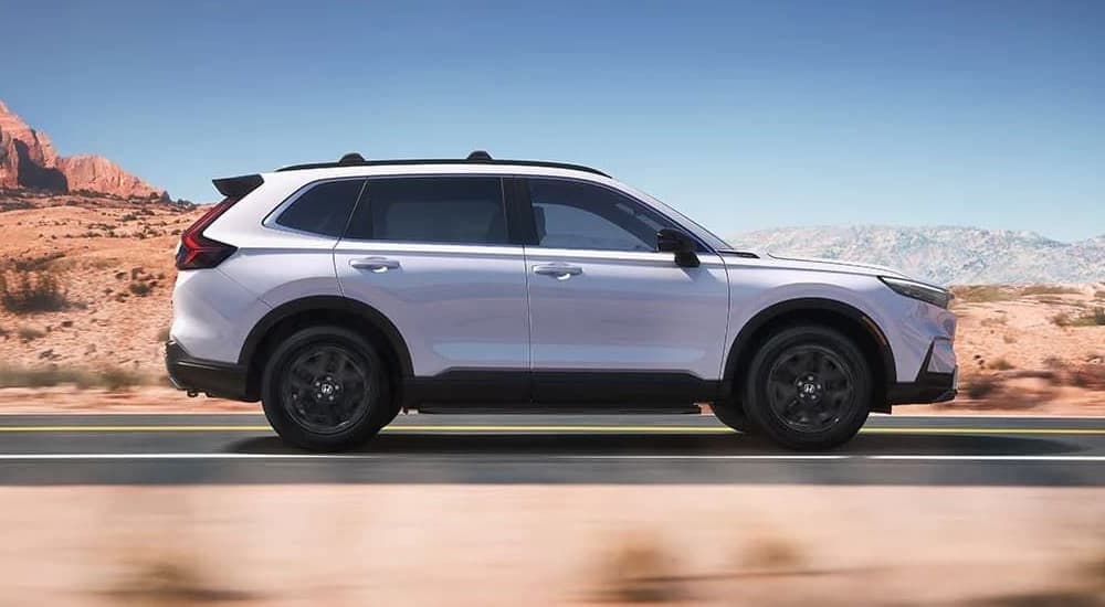 Side view of a white 2025 Honda CR-V Sport Touring Hybrid driving on a road.