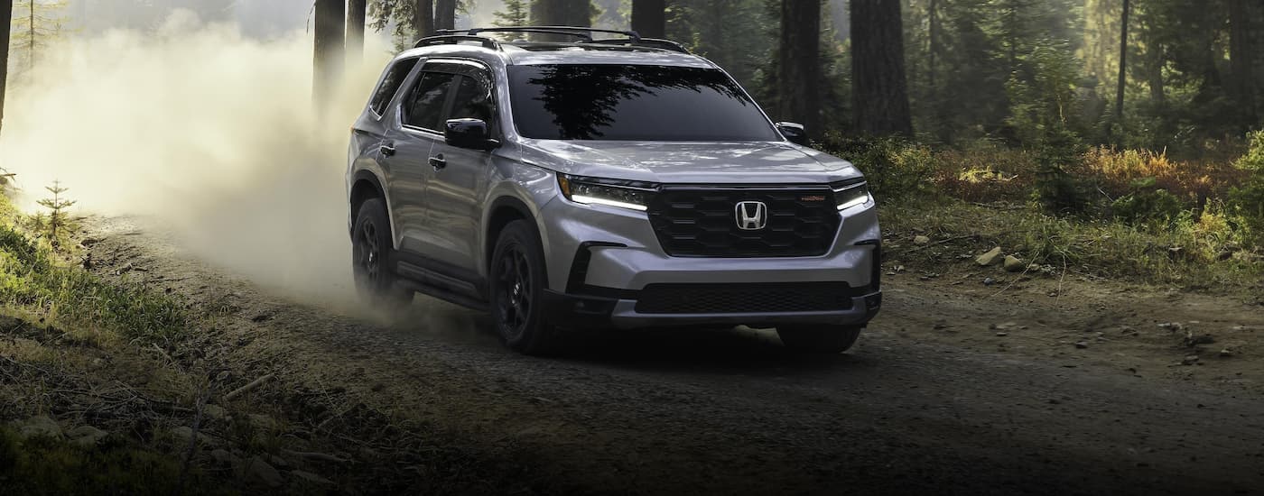 A grey 2025 Honda Pilot Trailsport driving on a dusty dirt road through the woods.
