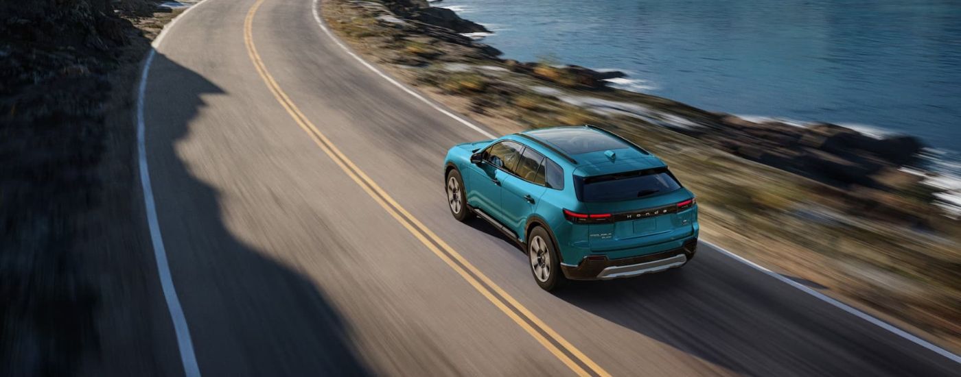 A teal 2024 Honda Prologue Elite is shown from the rear driving past a lake.