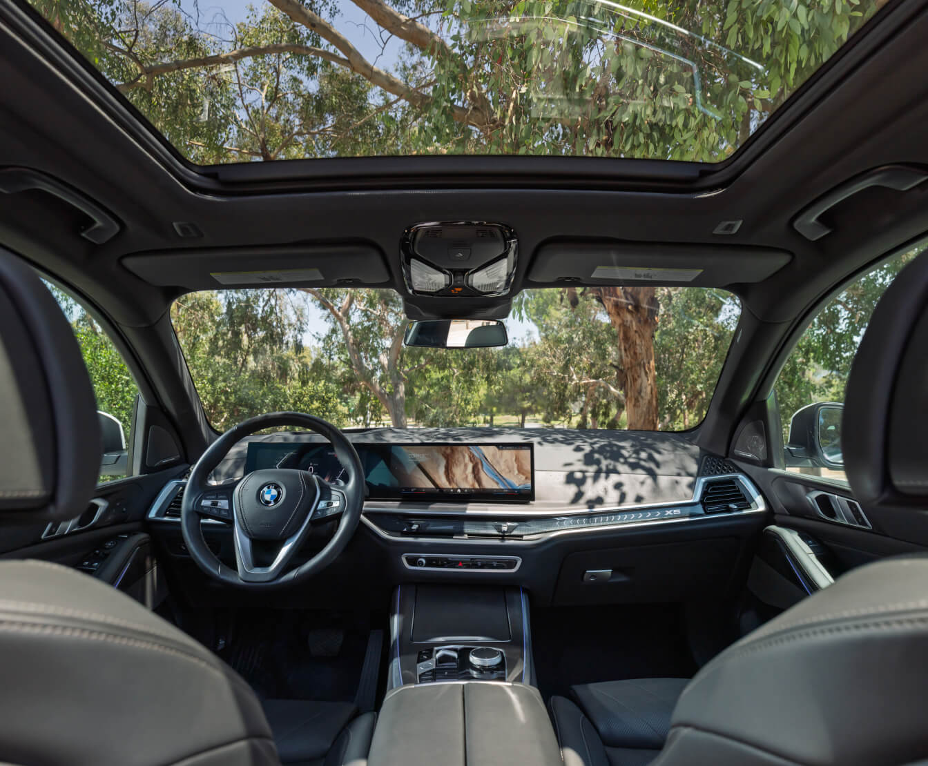 Interior front seat and dashboard of the 2026 BMW X5 with green trees showing outside the moon roof and windshield