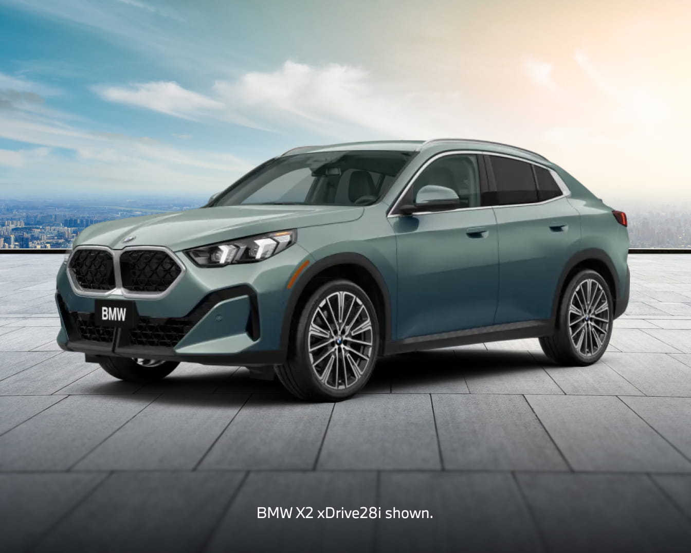 A green BMW X2 xDrive28i SUV shown from a front three-quarter angle on a paved plaza, with a city skyline in the background.