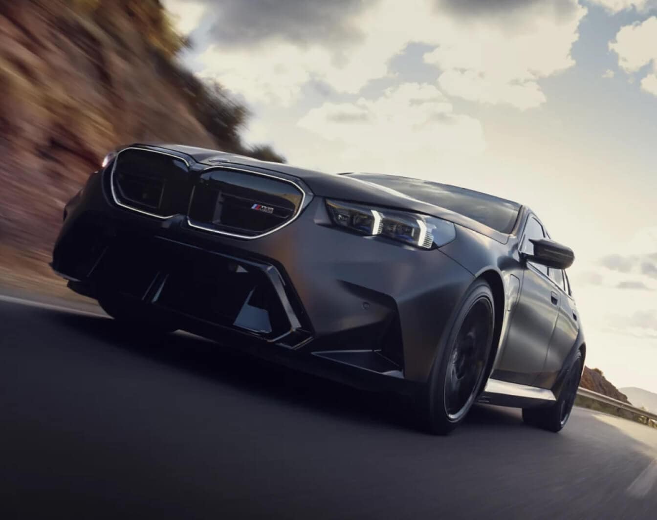 A front exterior low angle action shot of a dark gray BMW M5 driving down a highway.