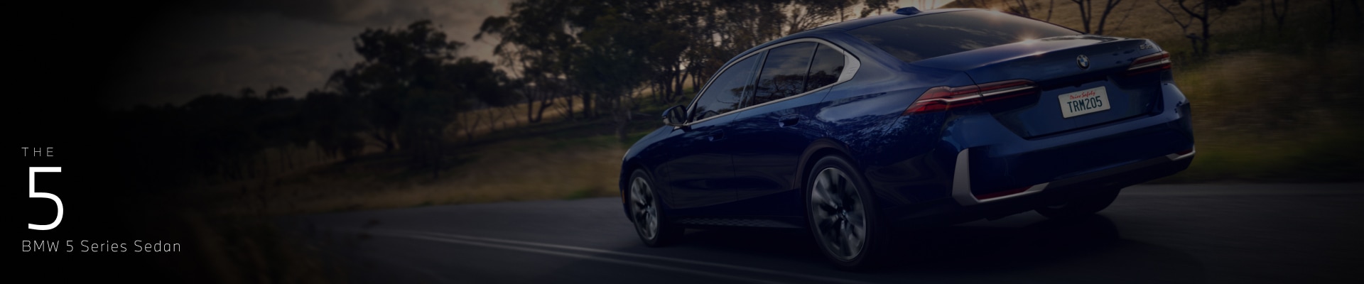 Three-quarter rearview shot of a 2026 BMW 5 Series in Blue driving with trees in the background