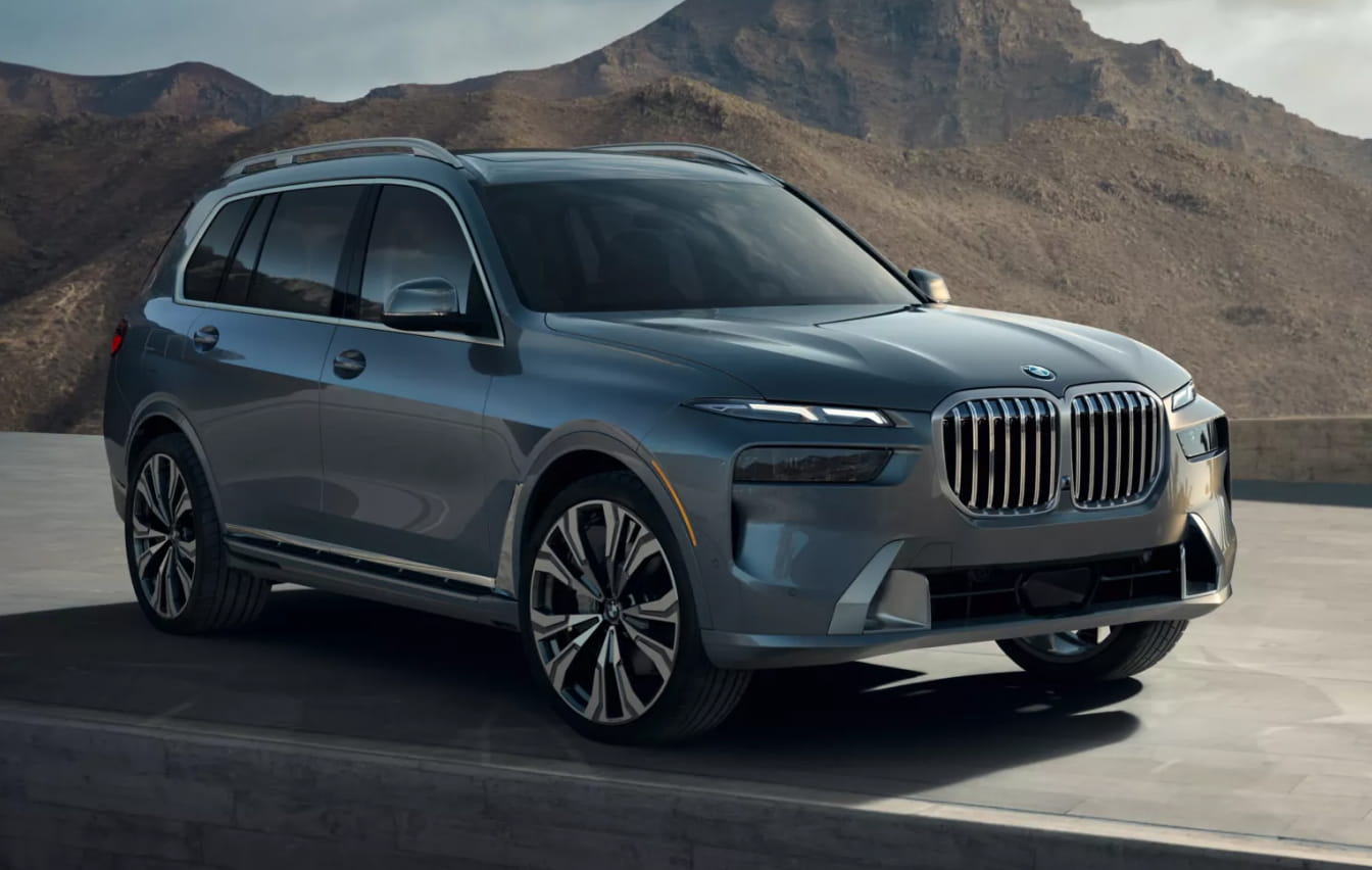 Blue-gray BMW X7 SUV parked on a paved terrace with rugged mountains in the background.