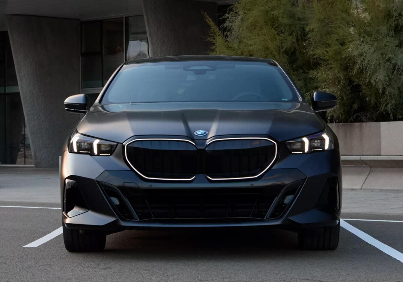 Front exterior view of a black 2027 BMW 550e parked in an office building lot showcasing the grill and headlights.