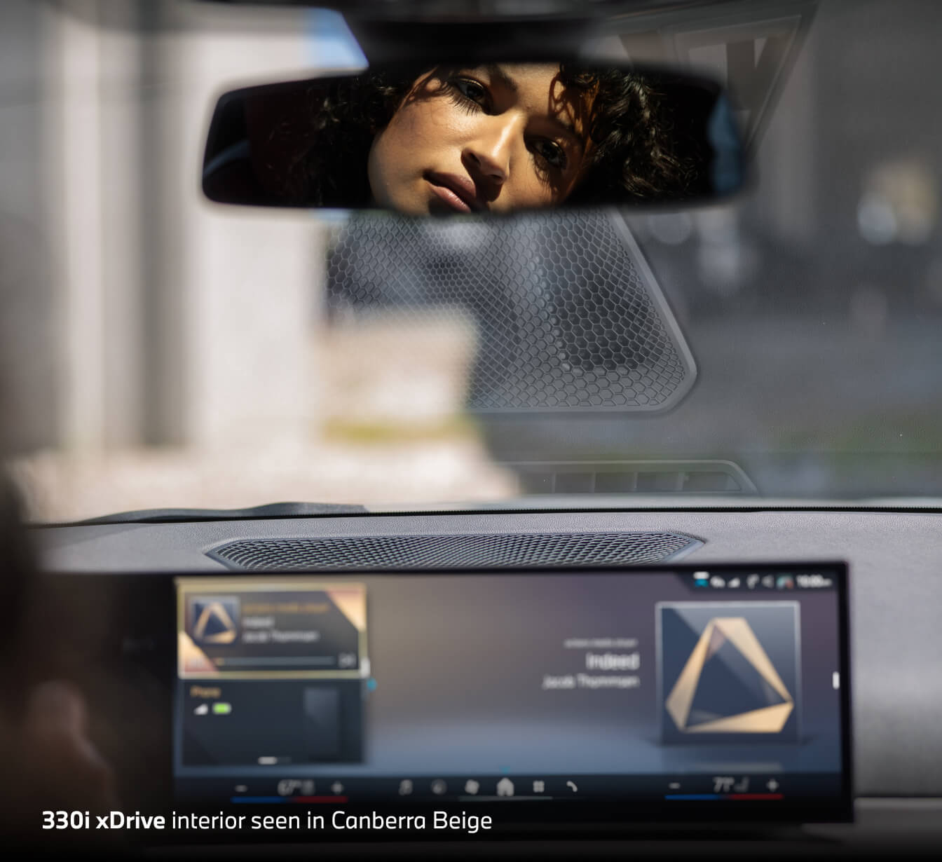 A woman's reflection in the rearview mirror above an infotainment screen. Words on bottom read 330i xDrive interior seen in Canberra Beige.