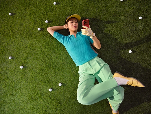  Person lying on their back on a lawn scattered with golf balls, looking at a smartphone.