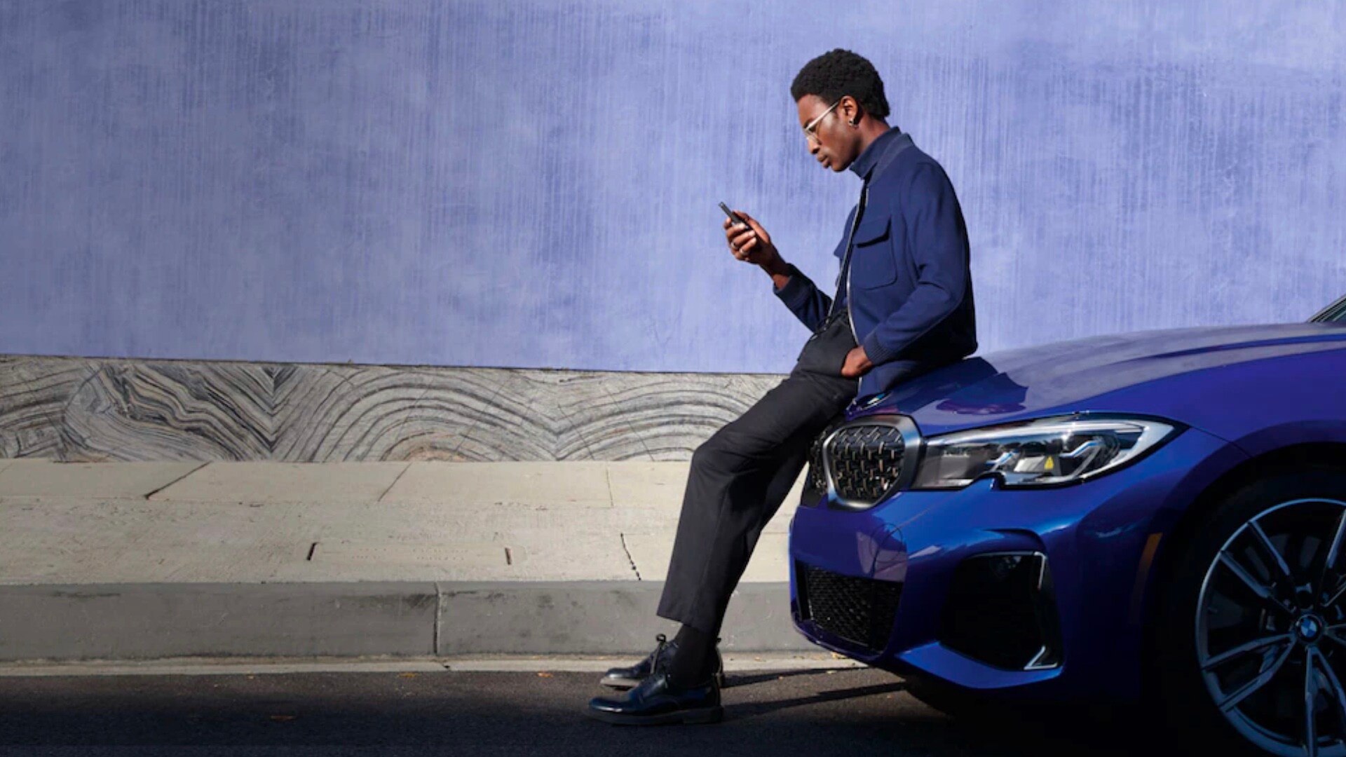 Man using smart phone sits on hood of a BMW