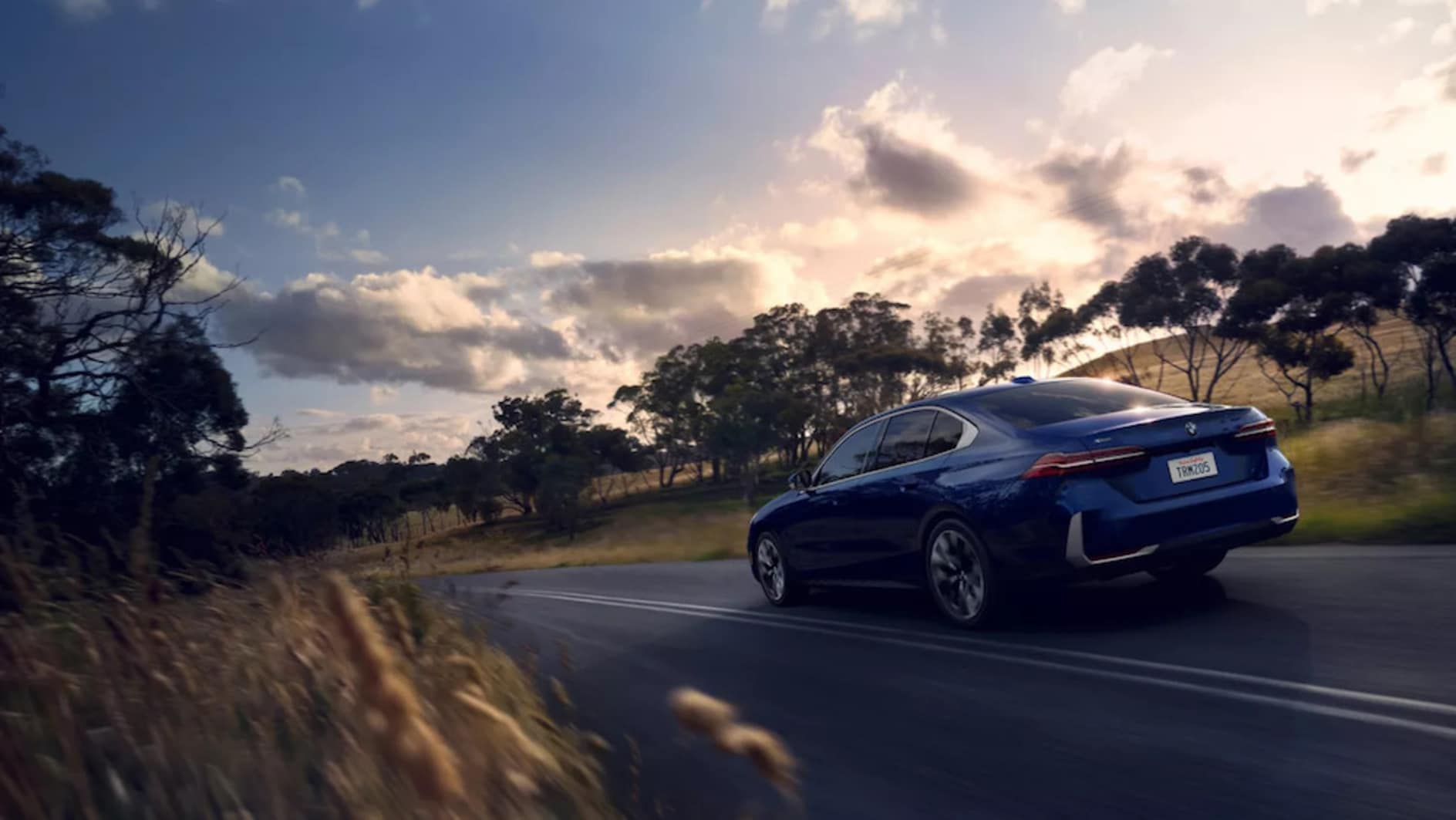 A blue 2026 BMW 530i sedan driving down a tree-lined road at sunset