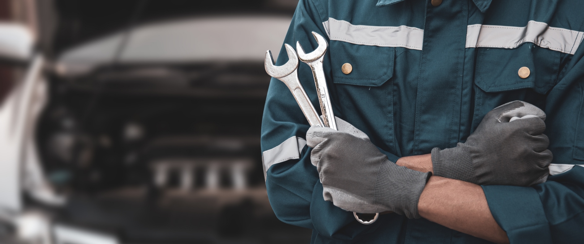 The torso of a person in a blue jumpsuit, holding two silver wrenches in a gloved hand, standing in front of an open engine of a car