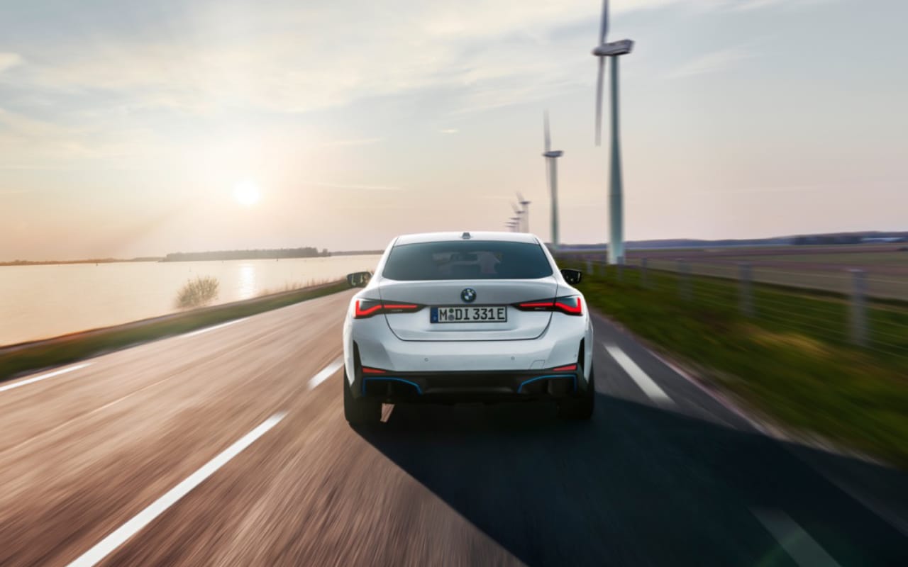 rear view of BMW i4 2023 at sunset on a road between a lake and a fieid of wind turbines