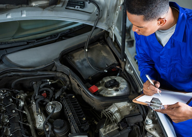 Everything You Need to Know about Your Car Battery at Bobby Rahal Acura | A mechanic in blue uniform inspecting the engine bay with a checklist