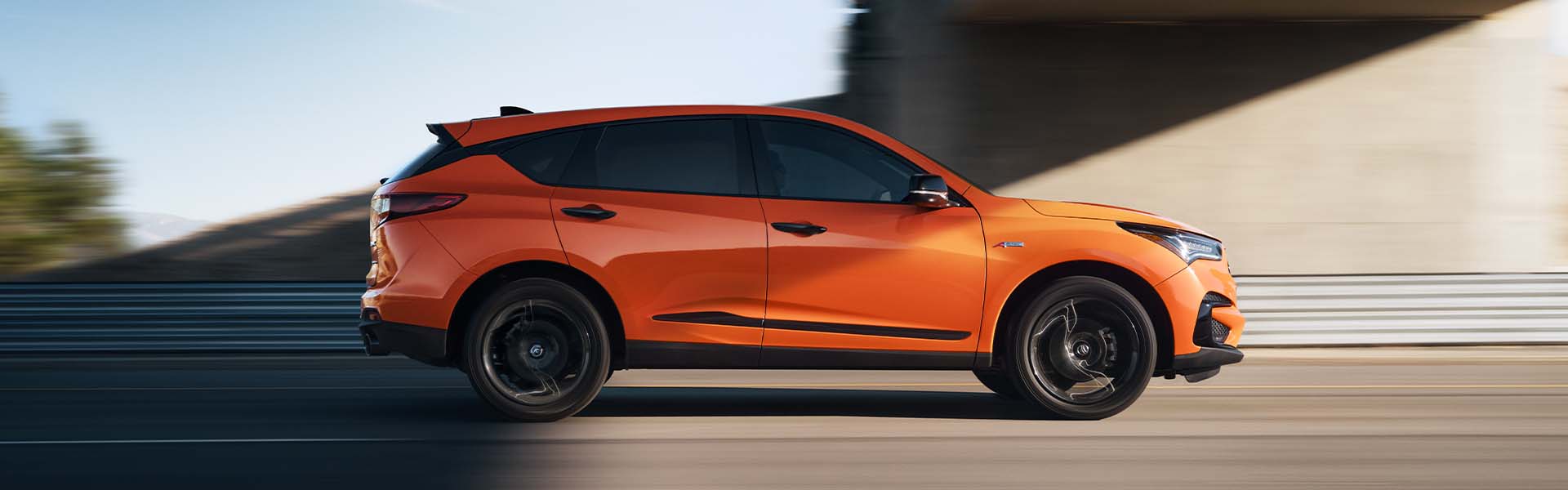 Compare the 2021 Acura RDX vs. the 2022 Audi Q5 at Bobby Rahal Acura | Profile View of Orange 2021 Acura RDX Driving Under Underpass on Highway at Sunset