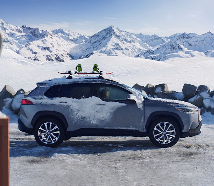 Model Features of the 2022 Toyota Corolla Cross at  Bobby Rahal Toyota | Silver 2022 Corolla Cross with Roof Rack Parked In Front of Snow Covered Mountains