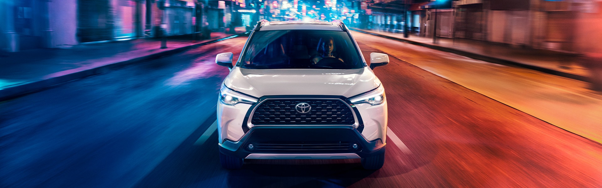 Model Features of the 2022 Toyota Corolla Cross at  Bobby Rahal Toyota | White 2022 Corolla Cross Driving Through Neon Lit City Streets at Night