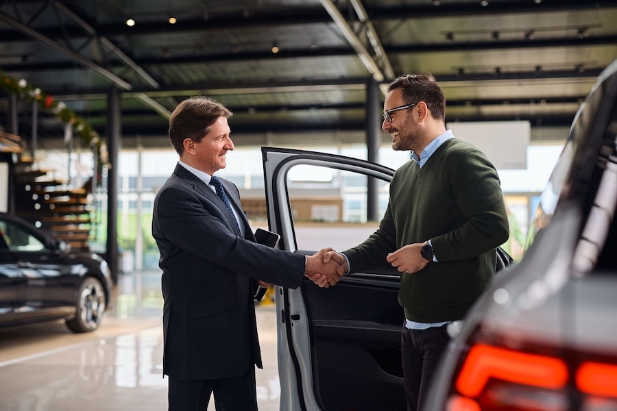 A car dealer and a customer shaking hands beside a vehicle inside a car dealership