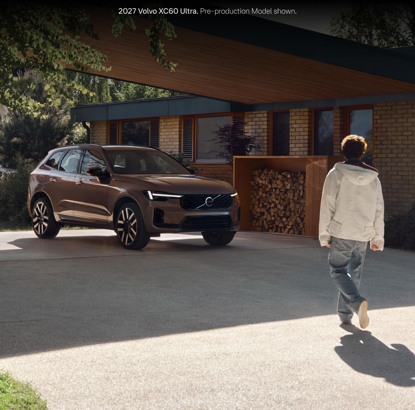 2027 Volvo XC60 Ultra in Heather Bronze parked on a modern residential driveway as a person in a hooded jacket walks past.