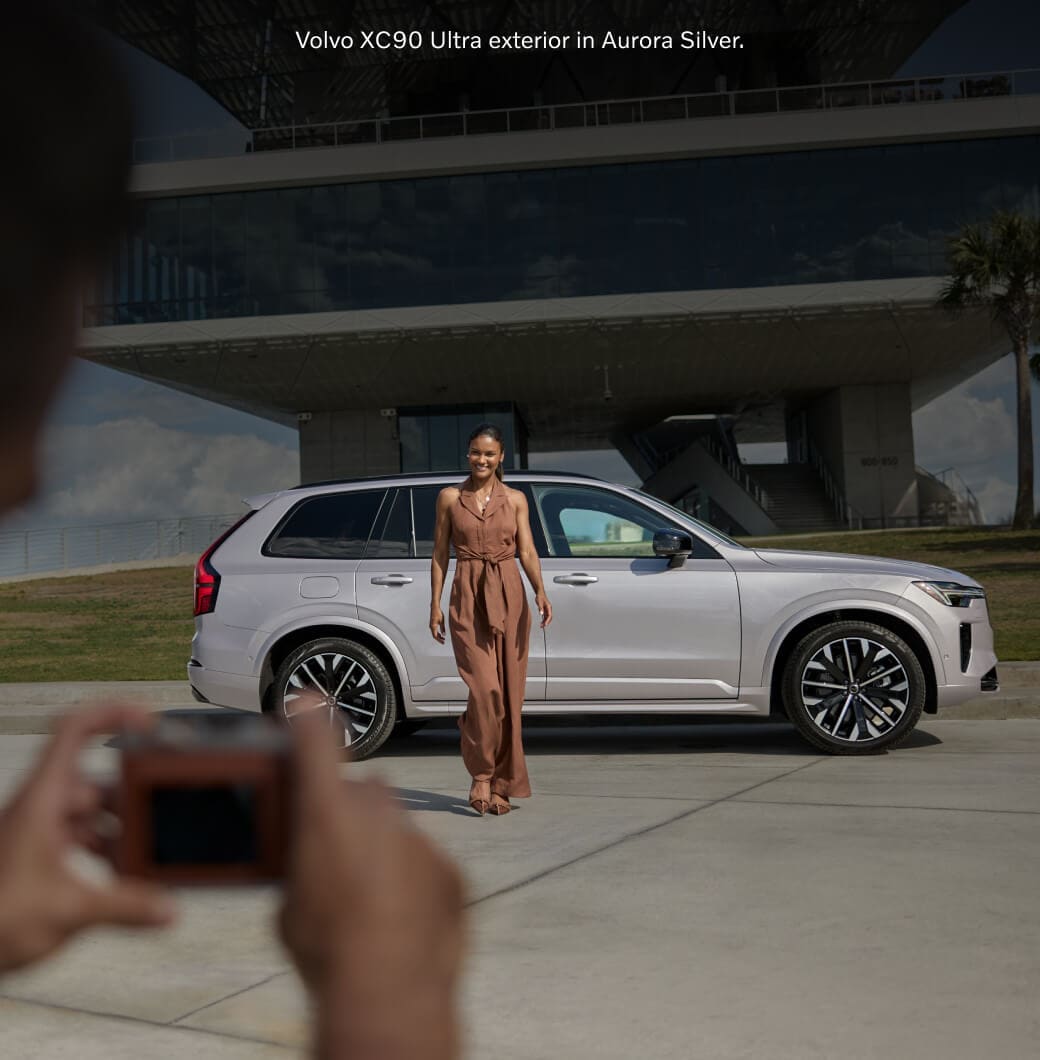 Side-profile view of an Aurora Silver Volvo XC90 Ultra with a woman posing in front as a photographer.