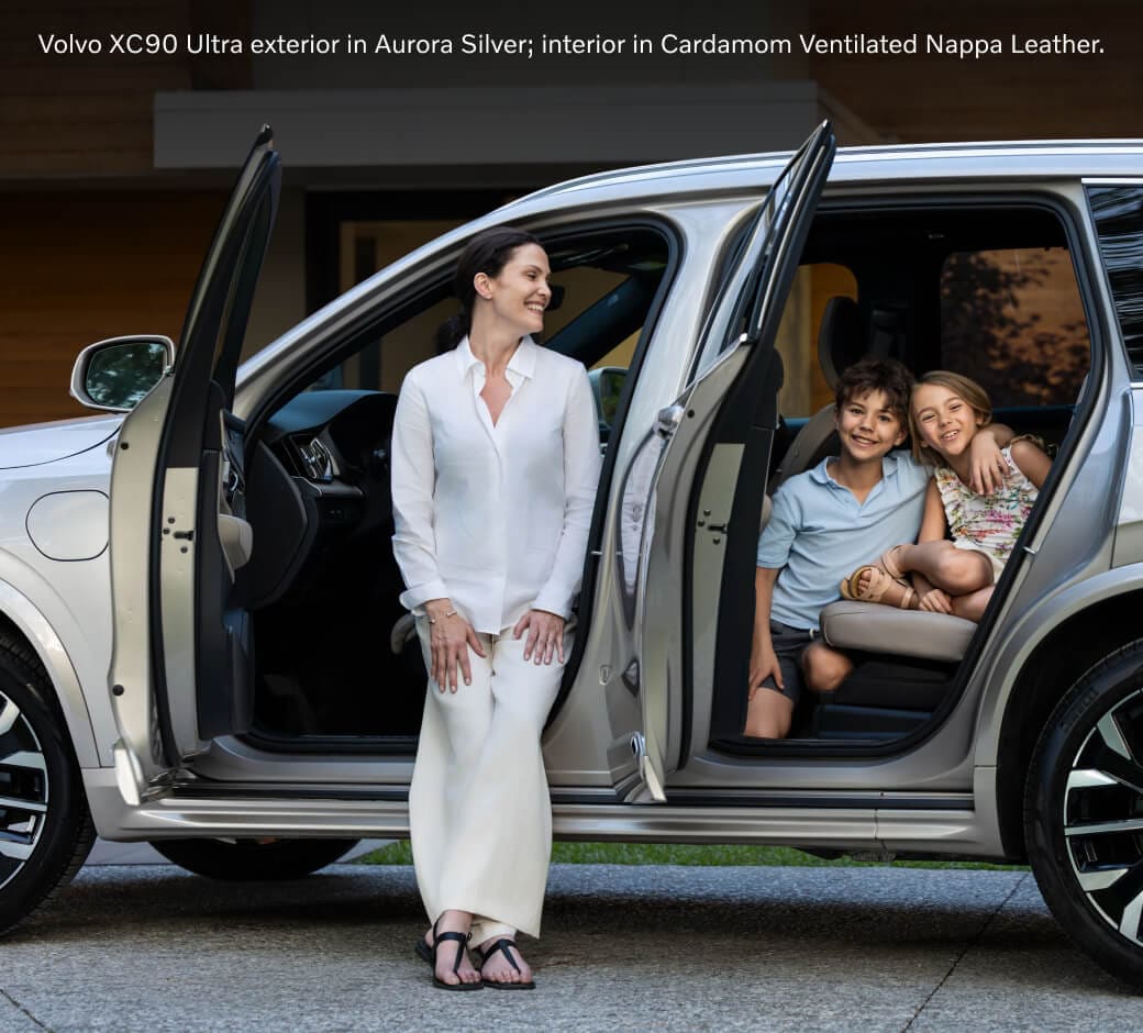 Side view of an Aurora Silver Volvo XC90 Ultra with open doors, a woman leaning on the sill, and children in Cardamom leather.