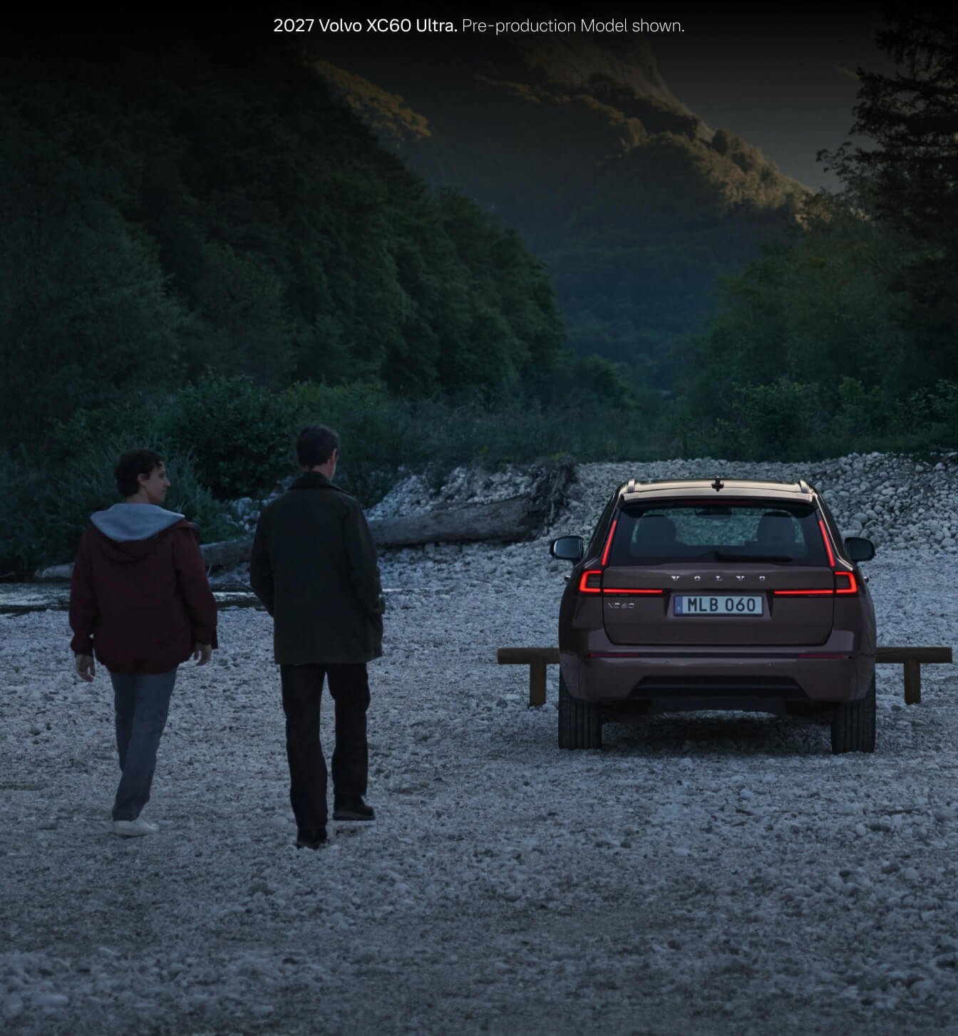 2027 Volvo XC60 Ultra in Heather Bronze parked on a rocky riverside bank at dusk with two people walking alongside.