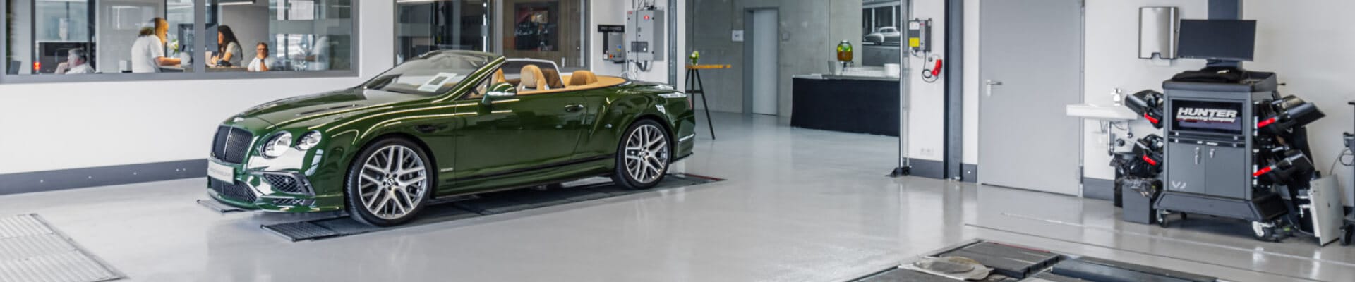 Green Bentley convertible with tan interior parked in a bright dealership service bay beside wheel alignment equipment.