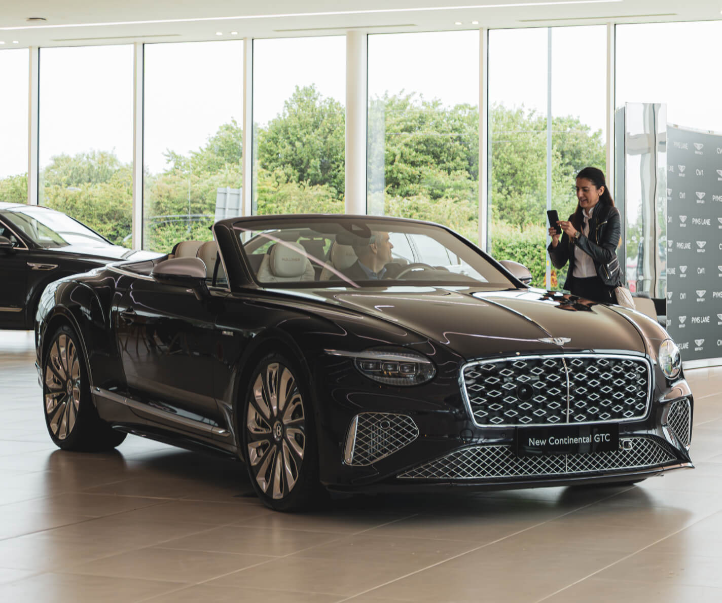 Three-quarter front view of a 2026 Bentley Continental GTC Speed in Beluga Black with an open top inside a showroom as a person takes a photo.
