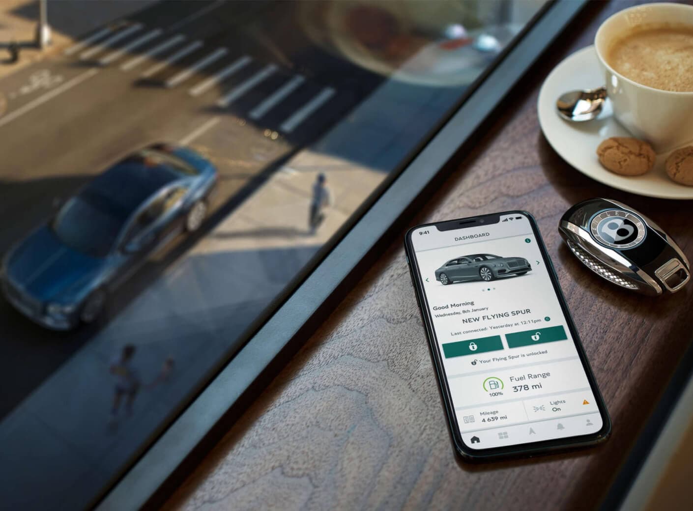 Top-down view of a smartphone showing the official My Bentley app beside a key fob on a café table.