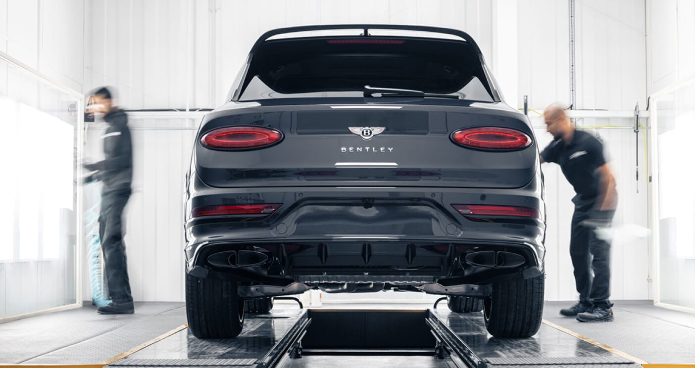 Rear view of dark grey Bentley Bentayga SUV on lift with technicians in well lit service bay.