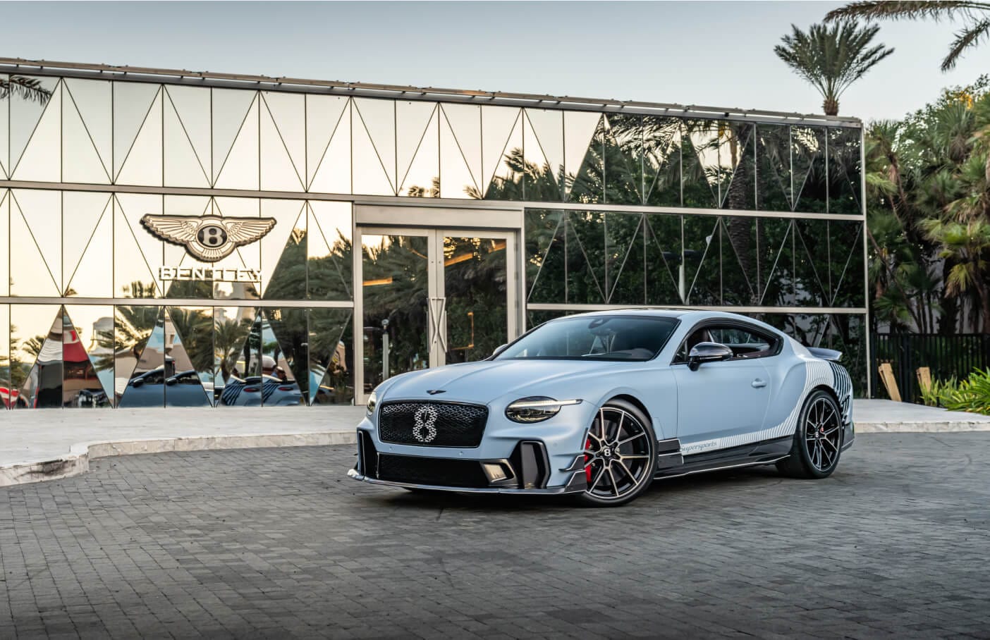 Front three-quarter view of light blue Bentley Continental GT Supersports in front of glass dealership.