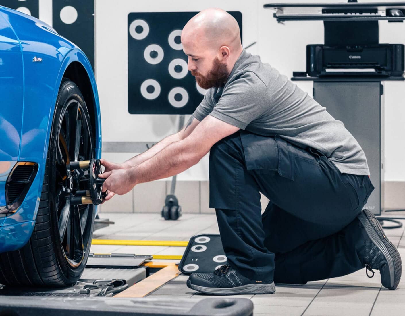 Side profile of a mechanic inspecting a wheel alignment on a blue Bentley in a service center.