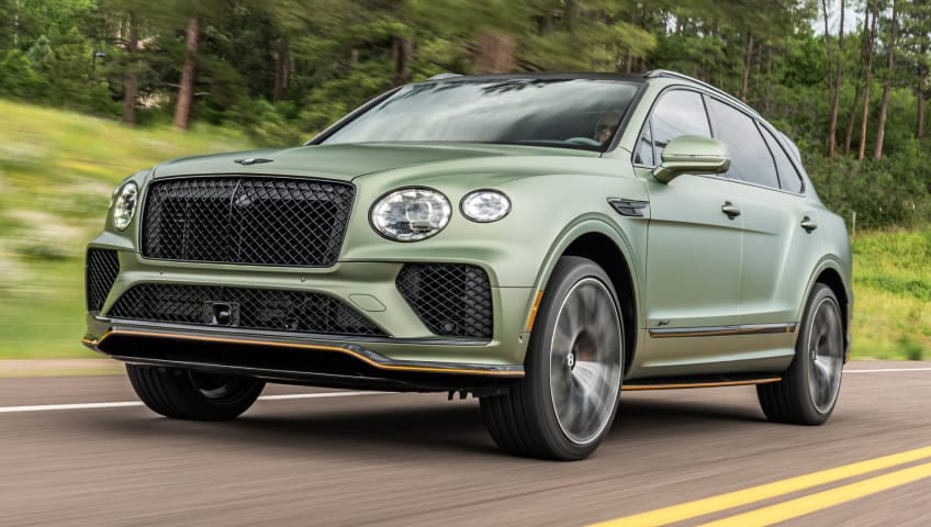 Green Bentley Bentayga driving on a tree-lined road.