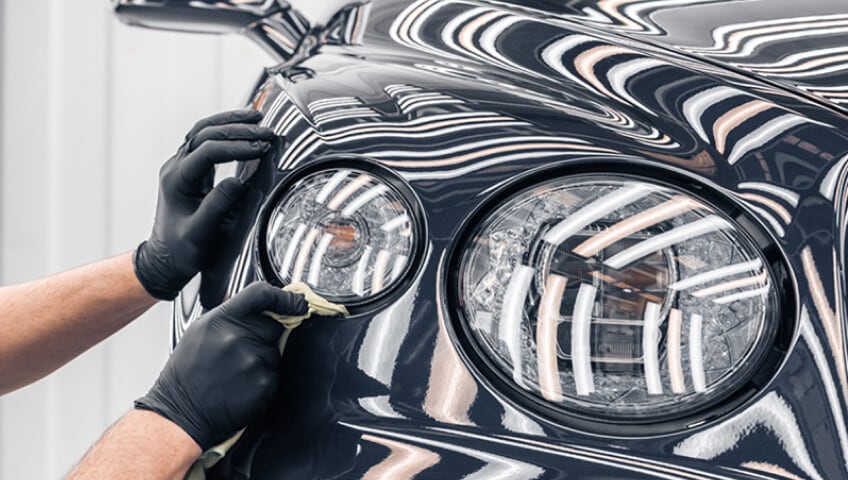 Front close-up view of technician polishing headlight on black Bentley in service bay.