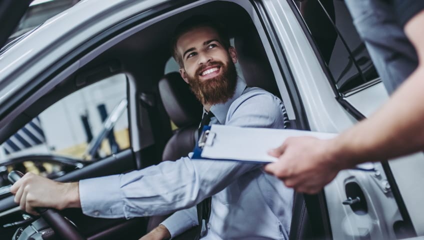 Service advisor handing paperwork to a smiling customer seated inside a vehicle.