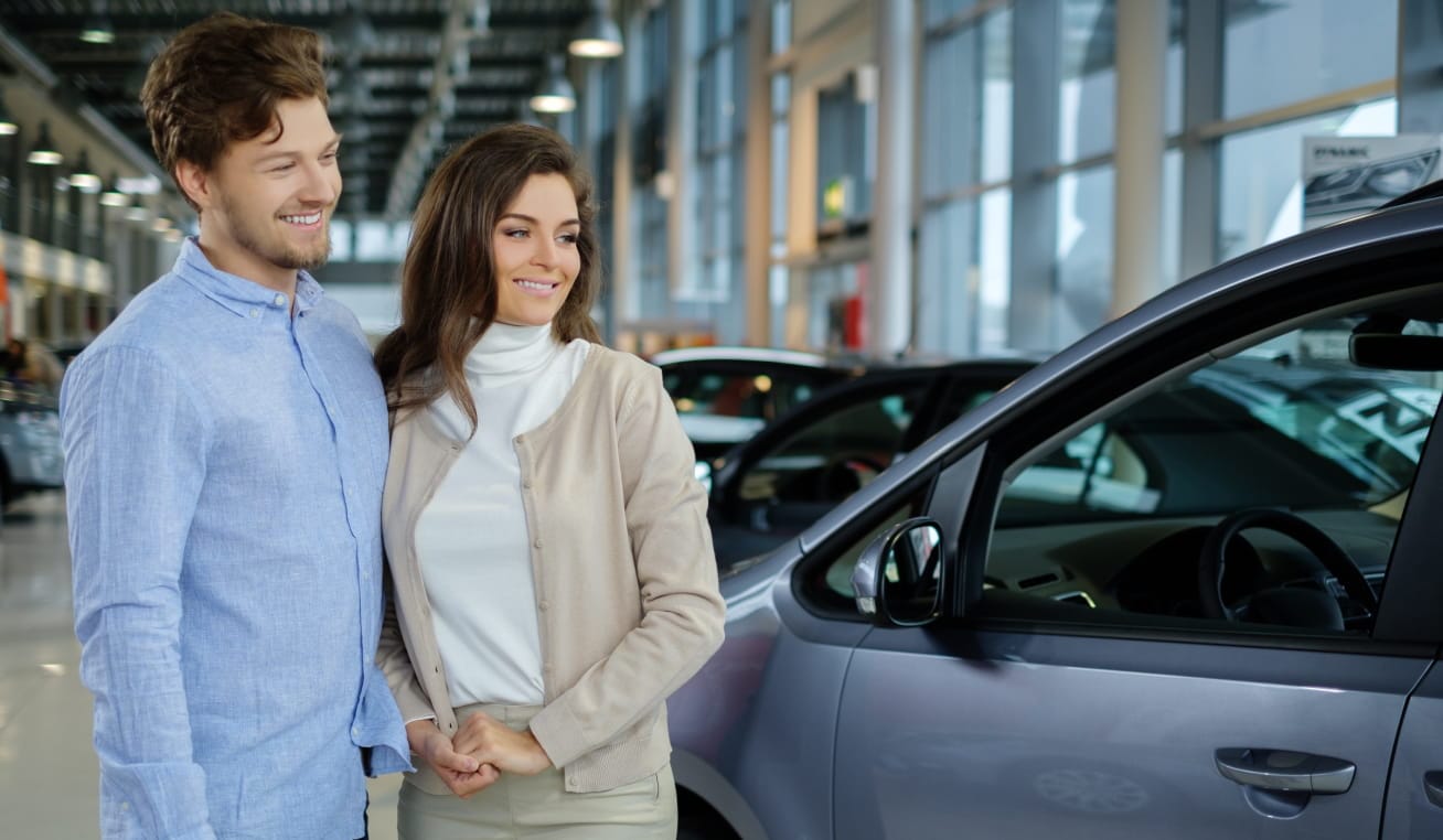 A smiling couple standing beside a vehicle inside a bright car dealership showroom.