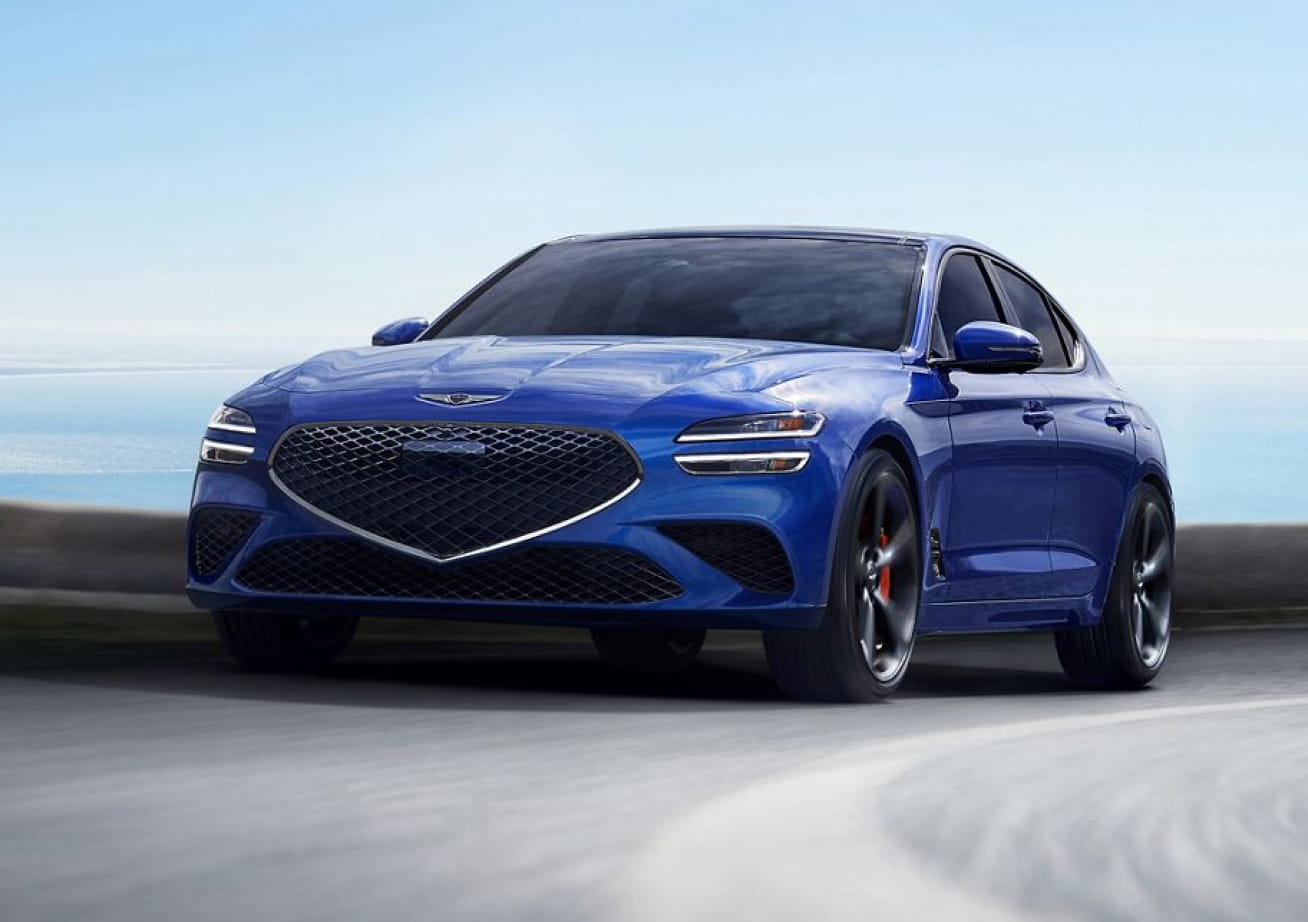 A blue Genesis sedan driving on a paved road, shown from a front three-quarter angle with a blurred roadway and open sky in the background.