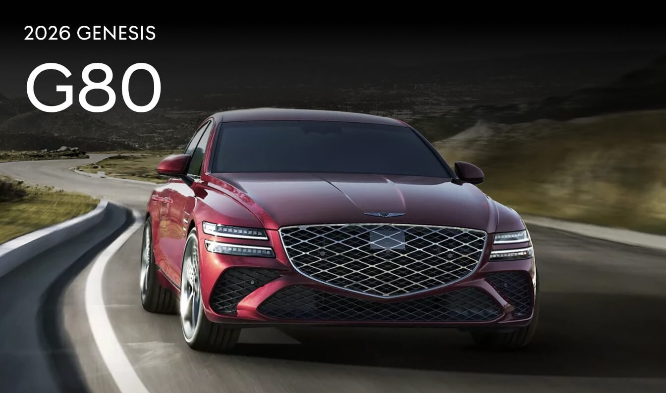 A front perspective action shot of a dark red 2026 Genesis G80 driving down a remote highway.