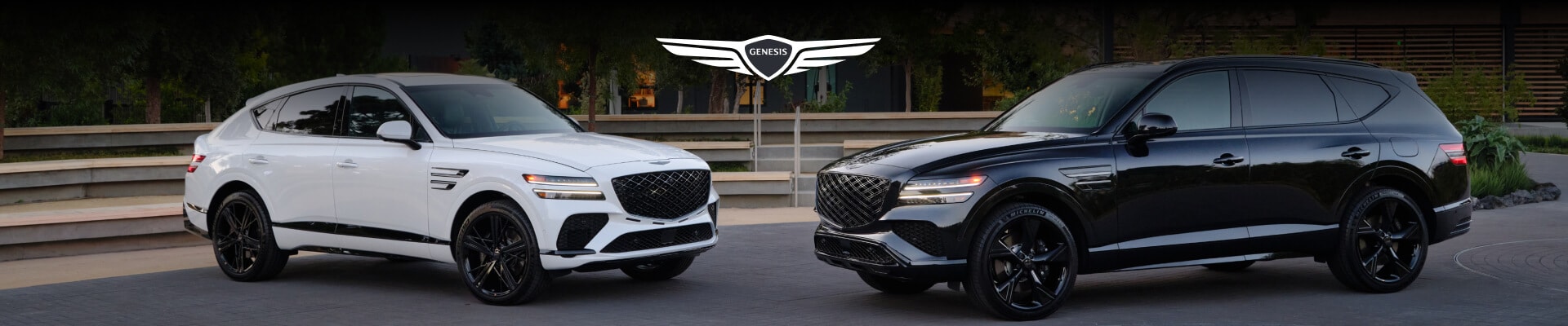 Three-quarter view of a white 2026 Genesis GV80 Coupe and black GV80 outside a modern dealership.