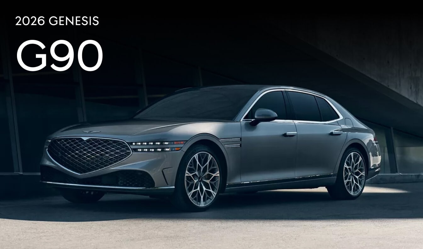 An exterior three-quarter view of a silver 2026 Genesis G90 parked in a shaded concrete parking lot.