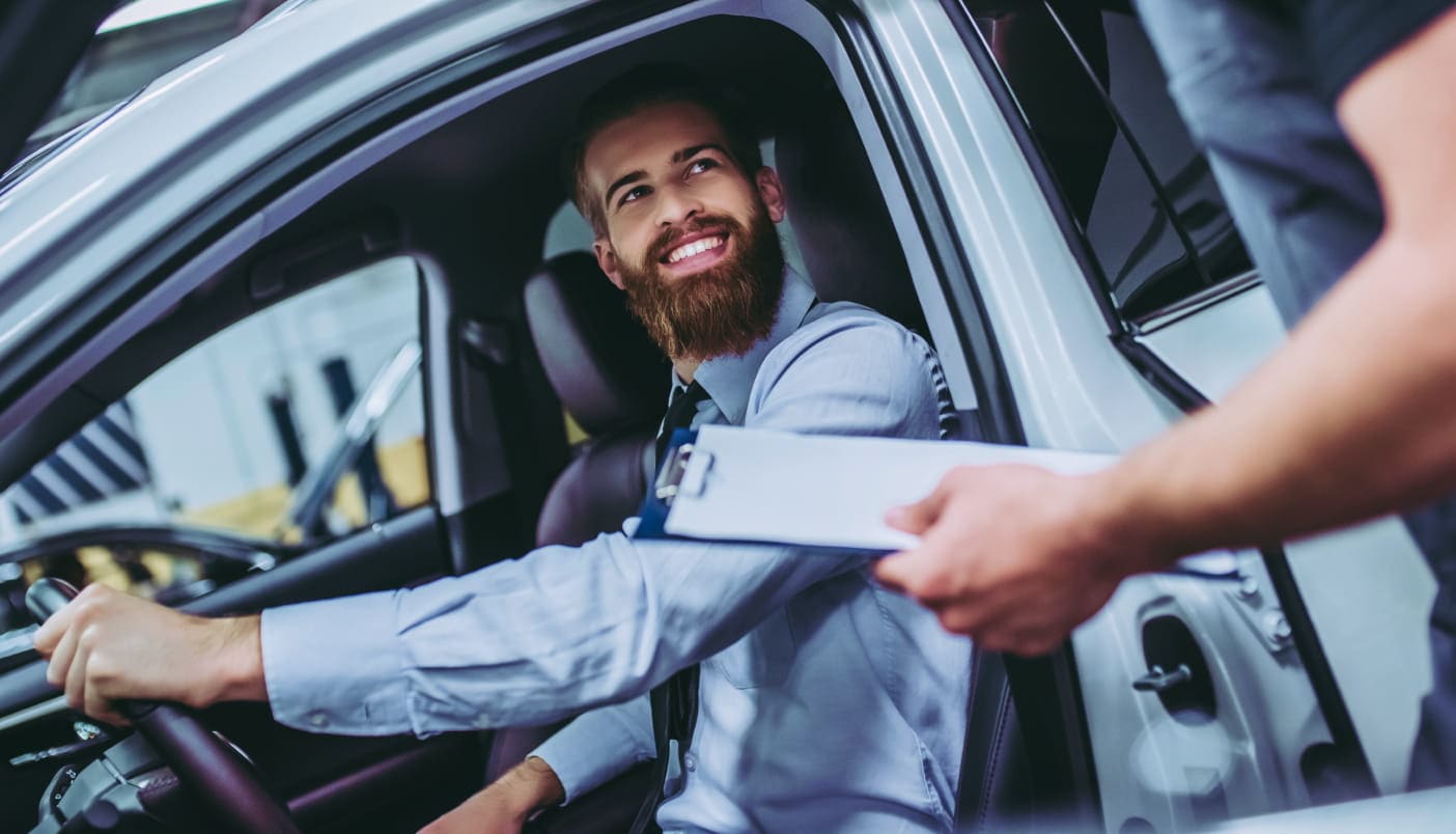Smiling customer seated in a vehicle while speaking with a service advisor holding a clipboard.