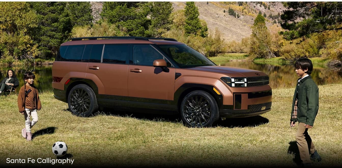 Side view of a copper 2027 Hyundai Santa Fe Calligraphy parked in a grassy clearing with two children playing soccer nearby.