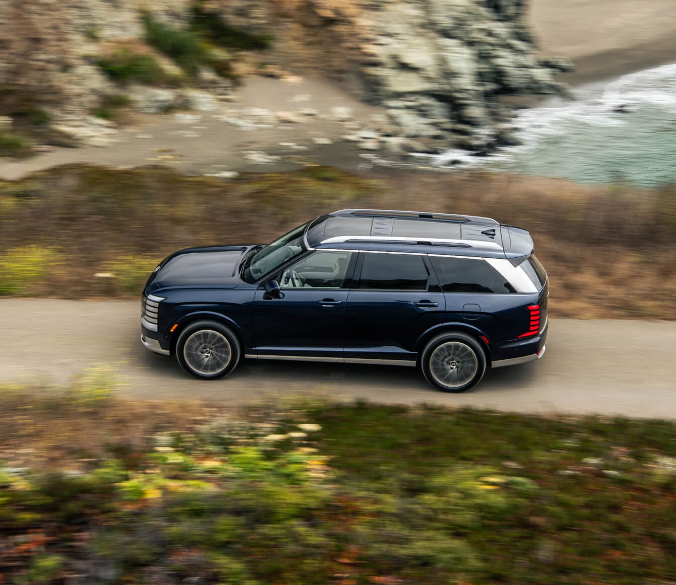High-angle side view of a dark blue Hyundai Palisade Calligraphy driving on a coastal road.